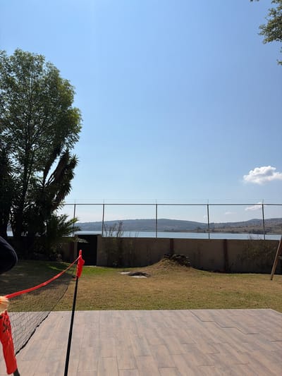 The image presents an outdoor scene in El Oasis Valsequillo, Mexico, under a clear sky with a single cloud. A chain-link fence runs across the mid-ground, behind which is a view of terrain and water, possibly a lake or reservoir. In the foreground, there are trees and vegetation. A section of tiled patio or walkway is visible on the right side. A badminton net is partially visible, with its poles and orange markers extending from the left into the patio area. The shot appears to be taken from a slightly elevated position. The overall environment suggests a residential or recreational area with a view.