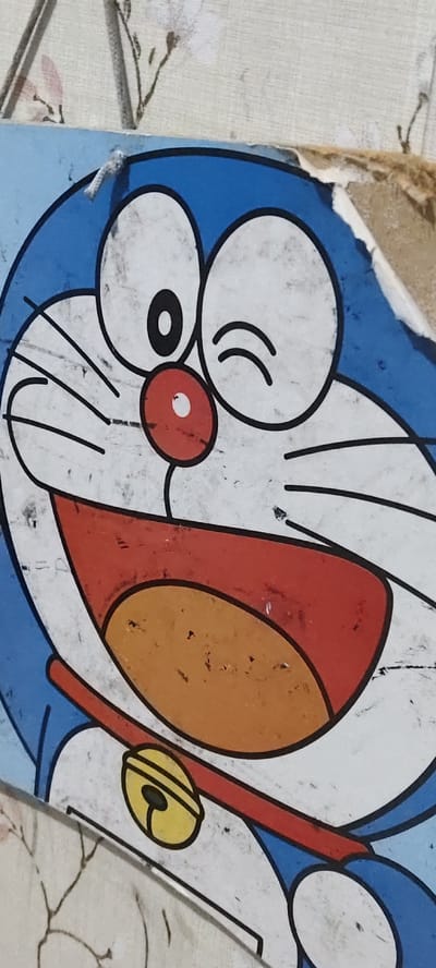 A worn illustration of the cartoon character Doraemon is depicted in a close-up, forensic manner, located in Kawungluwuk, Indonesia. The blue and white character's face and upper torso are visible, featuring a red circular nose and black whiskers. The right eye (viewer's left) shows a single black pupil, while the left eye (viewer's right) is winking. A wide, open mouth with a red outline reveals an orange interior. A red collar with a yellow, black-outlined bell is present on Doraemon's chest. The illustration exhibits extensive surface abrasion, scratches, and accumulated dirt. Its top and right edges are significantly damaged, peeling, and frayed, exposing a brown substrate. The background, visible above the illustration, is a light-colored, textured surface, possibly a wall, adorned with faint, stylized botanical patterns. A white thread or fiber is observed near the top-left edge of the illustration.