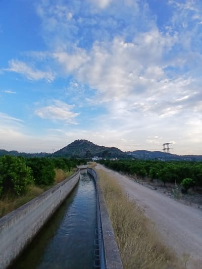 The image depicts a canal of water flowing through a rural landscape in Senyera, Spain. The canal, constructed with concrete walls, is bordered by dry grass on one side and lush green trees on the other. In the distance, a prominent hill with scattered buildings rises against a blue sky filled with white clouds. Power lines and a large electricity pylon are visible further in the background, suggesting the presence of infrastructure. The scene appears to be a natural or agricultural area, possibly an irrigation channel feeding the surrounding vegetation.