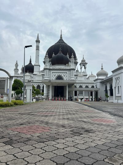 The image captures the grand architecture of the Zahir Mosque, a prominent landmark in Alor Setar, Malaysia. The mosque features a striking white facade with intricate gray detailing and is crowned by several dark domes. A tall, slender minaret stands to the left of the main structure. In front of the mosque, a wide paved walkway composed of interlocking gray and red patterned tiles leads towards the entrance. Several orange traffic cones are placed at intervals along the walkway, and a street lamp is visible on the left. The sky is overcast with gray clouds. Two motorcycles are parked on the right side of the paved area in front of the mosque's extended structures, and a person is seated on one of them. The overall scene suggests a place of worship, likely on a cloudy day, with some activity around the entrance.