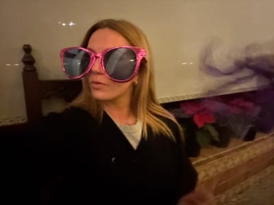 A person with long blonde hair wears oversized, pink-framed sunglasses. They are positioned upside down in the image. The person is dressed in a black top with a white collar. A box with purple, red, and green tulle fabric sits to the left. The scene includes a cream-colored surface, possibly a table or floor, and a dark wooden chair leg in the background. The lighting appears indoor and ambient.