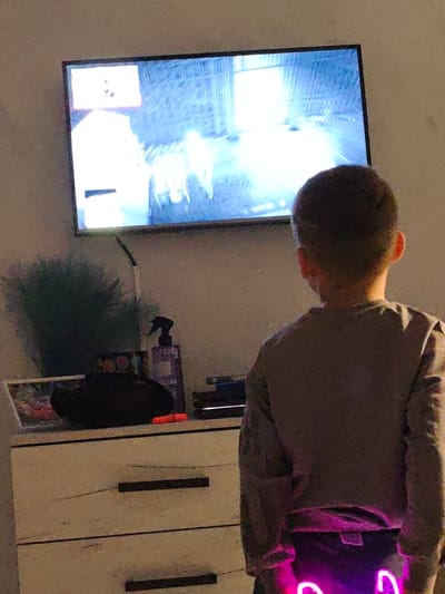 A young individual, viewed from the back, stands facing a wall-mounted flat-screen television in a room located in Opaka, Bulgaria. The individual, appearing to be a child with short, dark hair, wears a light-colored long-sleeved shirt and dark lower garment with two glowing pink and purple light patterns on the posterior. The television screen displays a brightly illuminated scene, consistent with a video game, featuring light-colored, possibly humanoid, figures or objects within a structured virtual environment, predominantly in shades of blue and white, with some user interface elements visible in the upper left corner. To the left of the scene, a white two-drawer dresser with dark horizontal handles holds a dark green fluffy object, a spray bottle with a black trigger, a colorful picture frame, an oblong dark object, an orange Nintendo Switch Joy-Con, and stacked dark media or electronics. The room is dimly lit, with the television screen providing the primary light source for the foreground and central area.