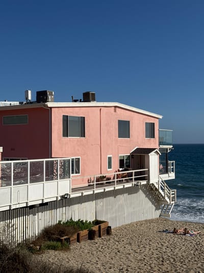 A salmon-pink stucco residential building in Malibu, United States, stands on a sandy beach under a clear, deep blue sky. The two-story structure features multiple dark-paned windows and a flat roof equipped with HVAC units, vents, and a white cellular antenna. A white wooden deck, supported by a vertical slatted foundation, extends from the house, partially shaded, with a section incorporating white-framed patterned glass panels on the left. A small covered entry structure leads to this deck, and a glass-railed balcony is cantilevered over the Pacific Ocean on the far right. White wooden stairs descend from the deck to the light brown sand. Two individuals are seated in lounge chairs on the main deck, while two other individuals are lying prone on towels on the beach below, sunbathing. The ocean exhibits gentle waves breaking on the shore. Dry vegetation and two wooden planter boxes are present in the sandy foreground.