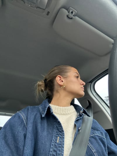 A young adult female is depicted in the front passenger seat of a vehicle, located in Saint-Cyr-sur-le-Rhône, France. She is seen from the chest up, facing right and looking out the side window, with her profile clearly visible. Her light brown hair is pulled back into a messy bun, secured with a black elastic tie. She has multiple piercings on her left ear, featuring a prominent gold-toned, floral or clustered earring on the lobe and several smaller studs along the helix. She wears a cream-colored, ribbed knit sweater beneath an open, dark blue denim button-up shirt or jacket. A dark-colored seatbelt is fastened across her chest and right shoulder. The vehicle's interior includes a light gray headliner, a light gray sun visor, and an overhead console with a light fixture. The exterior environment visible through the window is indistinct and appears overcast or rainy. No other individuals or direct interactions are visible.