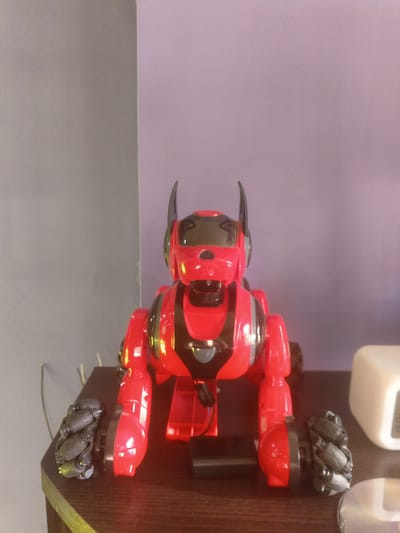 A red and black toy robot dog with articulated limbs and wheels is positioned centrally on a dark wooden surface. The robot has pointed ears, a dark visor for eyes, and visible wiring. To the left and right of the robot are its front wheels, which are dark grey and have a segmented, multi-wheeled design. A thin grey wire extends from the robot towards the left edge of the frame. Behind the robot, the background consists of a wall that transitions from a grey color on the left to a light purple color on the right. A white cubic object is visible on the right side of the frame, partially obscuring the purple wall. The scene appears to be indoors, potentially in a domestic setting. The location of Подымалово, Russia is not depicted or suggested by any elements within the image.