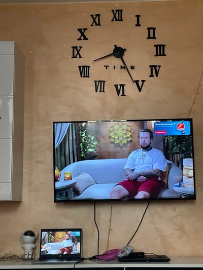 The image depicts an indoor living space. Dominating the wall is a large, black, Roman numeral clock. Below the clock, a flat-screen television displays a video of a man sitting on a couch. The man is wearing a white t-shirt and red shorts and has tattoos on his arms and neck. A laptop is positioned to the left of the television, also displaying the same video. A small, white astronaut-shaped lamp sits on the shelf next to the laptop. In the foreground, a black gaming console is visible. The overall environment appears to be a home in Russia, specifically within the residential complex "ЖК «Борисоглебское»".