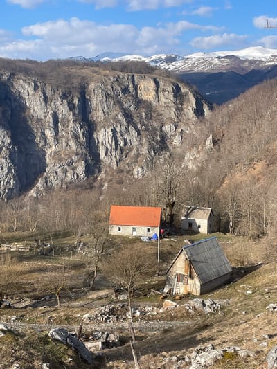 The image depicts a rural mountainous landscape in Mokro, Montenegro, under a clear blue sky with scattered white clouds. Distant snow-capped peaks rise above an extensive, steep grey rocky cliff face and densely forested hillsides primarily composed of bare deciduous trees. In the mid-ground, nestled on a sloping, partially cleared terrain, are three stone-built structures.
The most prominent building, positioned centrally, is a two-story stone house with a bright orange-red gabled roof, white-framed windows, and a white front door. To its right, two individuals are engaged in activity; one is crouched near a blue tarp on the ground, while another stands nearby. A vertical pole, possibly a utility or flag pole, stands in front of this house. Behind this structure, partially obscured by trees, is a smaller, older stone building with a darker, gabled roof.
In the lower right foreground, a third rustic stone building features a corrugated grey metal gabled roof and a wooden upper facade. A wooden ladder leans against its front. The immediate foreground consists of dry grass, scattered rocks, small bare trees, and visible fencing constructed from wooden posts and wire to the left of the central house. The ground around the structures exhibits signs of disarray, including scattered rocks, debris, and what appears to be stacked materials.