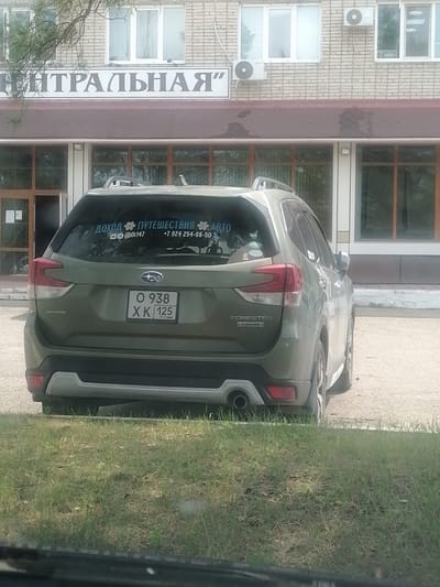 The image shows the rear of a green Subaru Forester parked on a paved area in front of a brick building. The building has multiple windows and a sign above the entrance that reads "ЦЕНТРАЛЬНАЯ". On the rear window of the car, there is a sticker with text and symbols, including what appears to be contact information. The license plate of the car is visible and reads "O 938 X K 125 RUS". To the left of the car, there is an entrance to the building, and to the right, more windows. The foreground is dominated by a strip of green grass. The scene appears to be outdoors, likely in a public or commercial area.