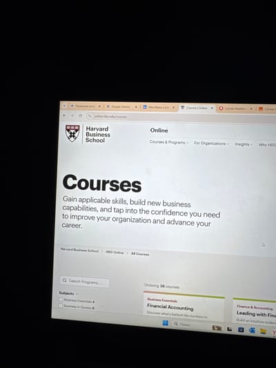 The image displays a close-up, angled view of a computer screen illuminated against a dark background, likely in a dimly lit room. The screen shows a web browser interface with multiple tabs open, including "online.hbs.edu/courses" currently active. Other visible tabs include titles like "Раскрытие ист...", "Google Gemini", "Alex Mazur | Li...", and "Cartela Vodafone".
The active webpage is the "Courses" section of the Harvard Business School's online platform. The page features the Harvard Business School logo and text "Harvard Business School" with "Online" as a subheading. A large heading reads "Courses," followed by descriptive text: "Gain applicable skills, build new business capabilities, and tap into the confidence you need to improve your organization and advance your career." Breadcrumb navigation "Harvard Business School > HBS Online > All Courses" is visible. A "Search Programs..." bar and a "Subjects" filter displaying "Business Essentials 4" and "Business in Society 6" are present. The page indicates "Showing 36 courses," with a partial view of two course listings: "Financial Accounting" under "Business Essentials" and "Leading with Finan" under "Finance & Accounting". A Windows taskbar showing a search icon labeled "Поиск" (Russian for "Search") and other application icons is partially visible at the bottom.
The image contains no discernible information to determine its specific physical location in the city of Ciorescu, Moldova. The depicted digital content is accessible globally.