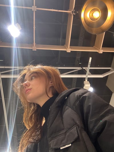 A woman with reddish-brown hair and fair skin is tilted to the right, wearing a black jacket. In the background is a room with what appears to be a wooden structure. A large, round light fixture illuminates the scene from above, and a smaller light source causes bright flares below. The room has an industrial or studio feel, with visible fixtures and rigging. The focus is on the woman's face and the details of her jacket and hair.