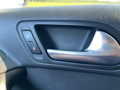 The image shows a close-up of a car's interior door panel. The focus is on the door handle and a button with a lock symbol. The door panel is made of black textured plastic, with a shiny silver door handle. To the left of the handle, there are two buttons. The top button has a symbol of a locked padlock, and the bottom button is plain. The overall condition appears to be clean and well-maintained. The background shows a blurred view of greenery and sky, suggesting the car is outdoors. There are no people visible in the image. The image is a static shot, with no discernible action taking place.