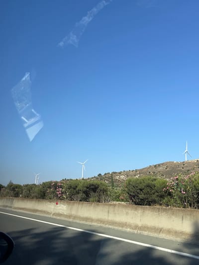 This image depicts a roadside view in Alethriko, Cyprus, under a clear blue sky, suggesting daytime. The scene is dominated by a road with a concrete barrier separating it from a hillside covered in vegetation and dotted with pink flowers. Several white wind turbines are visible on the hillside, indicating a location with renewable energy infrastructure. The reflection on what appears to be a window or car door shows geometric shapes and a distorted view of the sky and possibly clouds. There is no visible text in the image. The overall impression is one of a sunny day in a semi-rural or coastal area.