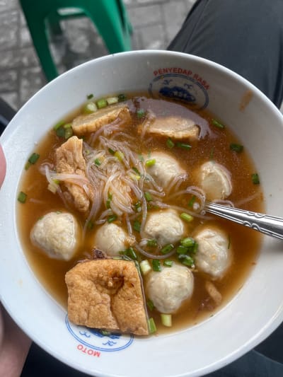In Kalijaten, Indonesia, a close-up, overhead shot captures a bowl of Indonesian meatball soup (bakso) served in a white ceramic bowl. The soup broth is a rich, reddish-brown hue, garnished with chopped green onions. Visible within the soup are several large, round, light-colored meatballs, thin, translucent noodles, and pieces of fried tofu. A silver spoon rests on the right side of the bowl, partially submerged in the broth, suggesting it's being prepared for consumption. The bowl itself features a blue and white emblem with text that reads "PENYEDAP RASA" and "MOTO" in a circular pattern, indicating the brand or origin. In the blurred background, a portion of a green plastic chair and a paved surface are discernible, hinting at an outdoor dining setting.