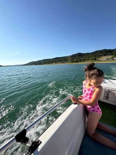 A young girl with dark hair in a ponytail, wearing a pink swimsuit with a strawberry pattern, leans over the edge of a white boat. She is looking down at the water, her eyes closed as if enjoying the sensation of the boat moving. The boat is on a body of water, possibly a lake or river, with green, tree-covered hills in the background. The sky is clear and blue, indicating a sunny day. The water is choppy, with white foam and ripples created by the boat's movement. A metal railing runs along the side of the boat, and the girl's hand is resting on it. There is a tattoo of a mermaid on her hand. The scene suggests a recreational activity, likely a boat ride on a warm day, possibly in the summer. The overall mood is peaceful and relaxed.