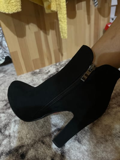 A black suede ankle boot with a zipper closure is worn on a person's left leg. The boot has a pointed toe and a thick heel. The person is standing on a grey patterned carpet. In the background, there is a light brown wooden surface with vertical planks, and some yellow fabric is visible at the top of the frame. The image was taken indoors. The specific location in the city of Kalininskaya, Russia cannot be determined from the visual information provided in the image.