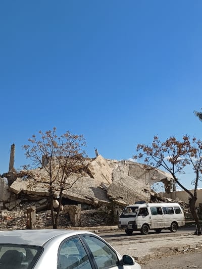 The image depicts a scene of extensive structural damage in Al-Qadam Municipality, Syria, under a clear blue sky. In the foreground, the silver roof and upper side window of a sedan are visible on the left. A light-colored, possibly white, passenger van or minibus is positioned on a paved road, facing left, with its front end partially obscured by a bare tree. The van appears to be in motion or recently stopped. Behind the road, a substantial structure is reduced to a large mound of concrete rubble and twisted rebar, indicating a catastrophic collapse. Scattered among the debris are several bare or sparsely leafed trees with brown foliage. No individuals are visibly present, nor are any direct interactions or specific actions definitively observable.