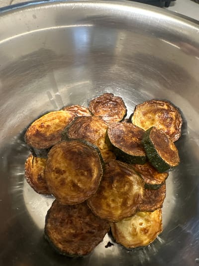 The image presents a close-up, overhead view of numerous circular slices of cooked vegetables, likely zucchini, gathered within a large, shiny metallic bowl. The vegetables, cut into thick rounds, display a spectrum of golden-brown to dark brown hues, indicative of pan-frying, roasting, or grilling. Many slices show a distinct, slightly crinkled or textured surface, with areas of deep caramelization alongside lighter, golden sections. The dark green skin of the zucchini is visible around the edges of several pieces. Some residual oil or moisture can be observed clinging to the vegetables and the bottom of the bowl. The metallic surface of the bowl reflects light, revealing slight scratches and a well-used appearance. The background is softly blurred, keeping the focus entirely on the prepared vegetables. The scene suggests an indoor setting, presumably a kitchen, where a meal component has just been cooked or is awaiting further preparation or serving. No individuals or visible text are present in the image, nor are there any discernible cues regarding the time of day or specific outdoor weather conditions, beyond the given context of Fulda, Germany.