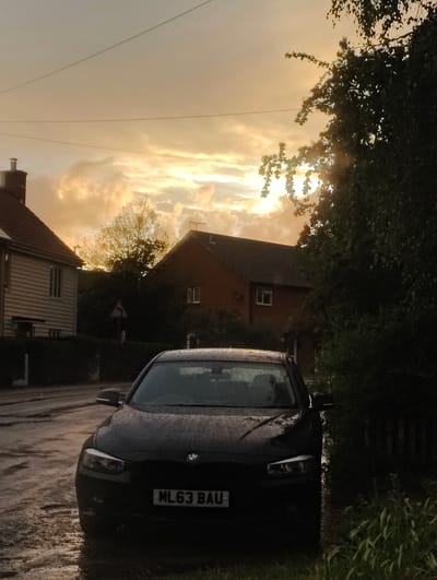 This is a low-angle, vertical, outdoor, eye-level shot of a black BMW sedan parked on a wet street in the city of Mid Suffolk, United Kingdom. The car is facing the camera and has water droplets on its windshield and hood. Its license plate reads "ML63 BAU". To the left of the car, there is a light-colored house with a dark roof and a brick chimney. To the right of the car, there is a dark green, dense bush and a wooden fence. The background shows two more houses, one a brick house with a dark tiled roof and the other a lighter colored house. The sky is filled with dramatic, orange-tinged clouds, suggesting a sunset or sunrise. The sun's rays are partially visible, shining through gaps in the clouds and trees. The street in front of the car is wet and reflects the light from the sky. A tree with yellowing leaves is visible behind the houses. The scene appears to be a residential street after a rainfall.