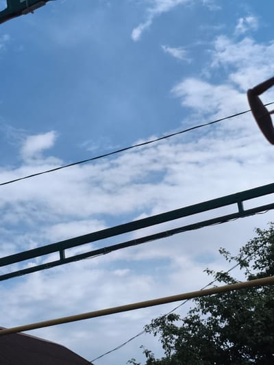 A low-angle, vertical shot captures a segment of the sky with scattered clouds and elements of urban infrastructure. In the foreground, a dark brown, ridged roof is partially visible at the bottom left, suggesting it is part of a building. Above the roof, a horizontal, yellow pipe stretches across the frame. Further up, two parallel, dark green, rectangular beams are positioned horizontally. These beams are secured by smaller white ties. A thin black wire runs diagonally across the upper portion of the image, and a partial view of a brown, curved object, possibly part of a swing set or similar outdoor equipment, is visible at the top right. Lush green foliage from a tree is present on the right side of the image, extending upwards from the bottom right corner. The sky above is a vibrant blue, with wispy white clouds. The scene appears to be an outdoor setting in an urban or suburban environment, potentially a backyard or park, in Barysaw, Belarus.