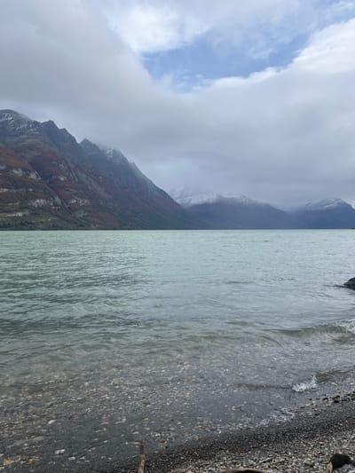 The image depicts a landscape near Lapataia, Argentina. On the left, there's a mountain range shrouded in clouds and mist, with some snow visible on the higher peaks. The mountain slopes are covered in vegetation, transitioning from green to reddish-brown. On the right, there's a body of water, possibly a lake or fjord, with a rocky shoreline covered in pebbles and stones. A small stick is visible on the right edge of the shoreline. The sky is overcast with a patch of blue peeking through the clouds on the left. No people, animals, or distinct landmarks are discernible in the image.