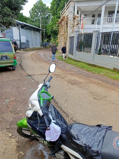 The image captures a rural or semi-urban street scene in Cimaung, Indonesia, under overcast daylight conditions. In the foreground, a green and white scooter is prominently parked, partially obscuring a puddle. A white plastic bag is tied to its handlebar, and its seat is covered with a black plastic sheet, with "JUPITER" visible on its side panel and "DORAEMON" faintly printed on the uncovered part of the seat.

The road itself is a mix of dirt and worn asphalt, showing several wet patches and small puddles, suggesting recent rain. To the left, a green van with the license plate "D 1081 CU" is parked. Behind it, a long, low building with a metal roof and light-colored walls is visible.

Further down the winding road, two men are walking away from the viewer on the right side. The man in front wears a dark jacket and blue jeans, while the one behind him wears a dark t-shirt and a cap. On the right side of the road, a two-story building is under construction, with extensive bamboo scaffolding covering its facade. In front of this building, a grey wall with black vertical metal bars serves as a fence. A small red and white flag, likely the Indonesian flag, is visible hanging from the balcony of the constructed part of the building. Lush green trees line both sides of the road and fill the background, indicating a verdant environment.