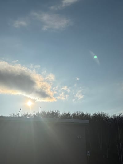 The image, oriented sideways, presents an outdoor scene dominated by a clear to partly cloudy sky on a bright, sunny day. When viewed with the sky at the top, a vast expanse of blue fills the frame, dotted with sparse white clouds. A prominent, elongated cloud formation stretches across the mid-section, appearing somewhat dark but with bright, sunlit edges in golden and white hues.
The sun is visible on the right side, partially obscured by clouds, casting intense rays of light downwards and outwards. A distinct, bright green lens flare is noticeable in the upper left portion of the sky.
At the bottom of the frame, a dense line of bare trees with intricate, dark branches forms a natural horizon, suggesting a colder season like winter or early spring. Below these trees, a light-colored, horizontal structure, possibly a building or a wall, extends across the width of the image, appearing somewhat reflective or brightly illuminated by the sun. Two dark lampposts or utility poles, with their lights off, rise from behind this structure into the tree line. In the lower right corner, above the structure and set against the trees, a blue square sign with a white "P" is visible, indicating a parking area.
The scene captures a moment of dramatic natural lighting, likely during the morning or late afternoon, in Ermolaevo, Russia. No people or specific activities are depicted; it's a static landscape.