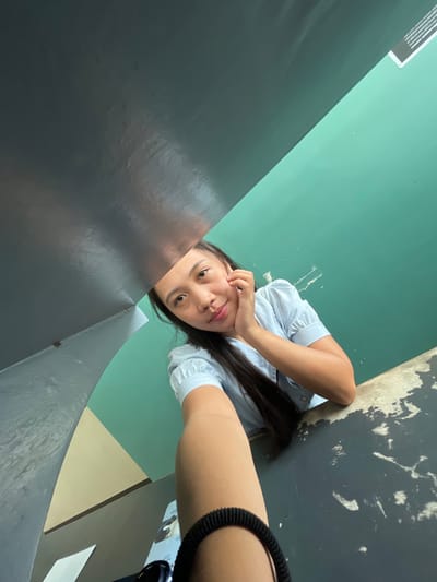 A young woman is captured in a selfie, looking towards the camera with a relaxed posture. She is leaning on a dark, textured surface, with her right hand touching her chin and her left arm resting on the surface. She is wearing a light blue collared shirt with short sleeves. The background consists of a teal-colored wall with some white marks and a beige triangular section on the left. A dark, glossy surface is overhead, partially obscuring the view. The lighting appears to be natural. There is a piece of paper with text visible on the dark surface in the foreground. This image was taken in Koronadal City, Philippines.