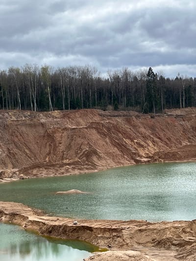 The image depicts a section of the Blue Lakes quarry located in Ivlevo, Russia. The quarry walls are layered and composed of brown soil. A body of water with a blue-green hue fills a portion of the quarry. A line of trees with bare branches borders the edge of the quarry on one side. The sky is overcast with gray clouds.