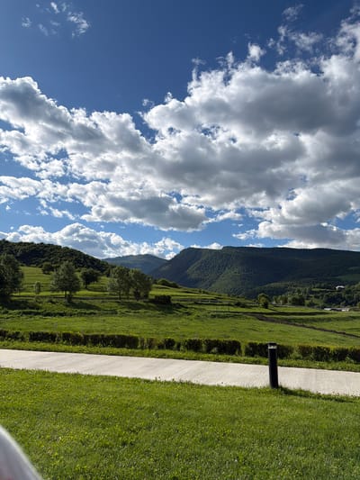 The image depicts a landscape with a clear blue sky adorned with white clouds on the left side, transitioning into a scene of green mountains, grassy fields, and trees on the right. The location is Chkalov, Armenia. A gray pathway cuts through the green grass and a black cylindrical object is positioned beside it. The grassy fields are manicured and bordered by hedges. The mountains are covered in lush green vegetation. The overall atmosphere suggests a serene and picturesque environment.
