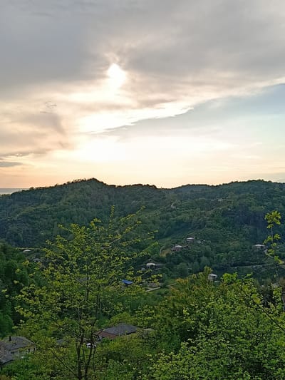 The image shows a hilly landscape in the city of Yuada-Gulrypsh, Abkhazia. Lush green vegetation covers the hills, with trees dominating the landscape. Several houses are scattered throughout the area. The sky is overcast with a break where the sun is visible.