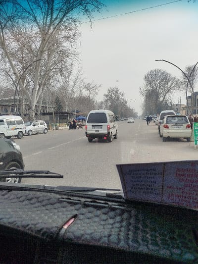 The image captures a street scene in Balykchi, Uzbekistan, viewed from the interior of a vehicle. The foreground features a dark, textured dashboard with two windshield wipers resting at the base of the windshield. A light-colored card with illegible text is positioned on the right side of the dashboard, partially obstructing the view. The upper part of the windshield displays a blue tint.
The midground shows a two-lane asphalt road with a dashed white centerline, indicating traffic flow. A white microvan, license plate 80 D 328 AB, is centered in the far lane, moving away from the viewer. On the right shoulder, a cream-colored hatchback, license plate 80 M 311 LG, is parked. On the left shoulder, a white microvan and a silver hatchback are parked. A group of approximately three to four individuals is gathered on the left side of the road near temporary market stalls covered by blue tarps.
The background is dominated by numerous large, bare deciduous trees with prominent light-colored bark, lining both sides of the avenue. Behind the trees on the left, architectural elements of buildings, including arches and columns, are discernible. Streetlights with curved poles are visible along the road. The sky is overcast and grey, indicating a cloudy day. Scattered pedestrians are visible on the sidewalks further down the street, which recedes into a slight atmospheric haze. The bare trees suggest the scene takes place during late autumn or winter.