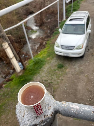 A white disposable paper cup, labeled 'COFFEE' in red, containing a reddish-brown liquid with a central swirl, rests on a weathered gray metal railing exhibiting chipped paint and rust. Below the railing, a white Lexus GX 470 SUV is parked on a dirt path, facing away from the viewer. The vehicle has a front license plate and a roof rack. Beneath the path, an embankment with sparse green grass and brown vegetation slopes down to a narrow stream with flowing water. The background features additional leafless trees and brush. No individuals are present. The precise location within the city of Lehvaz, Armenia, cannot be determined from the image.