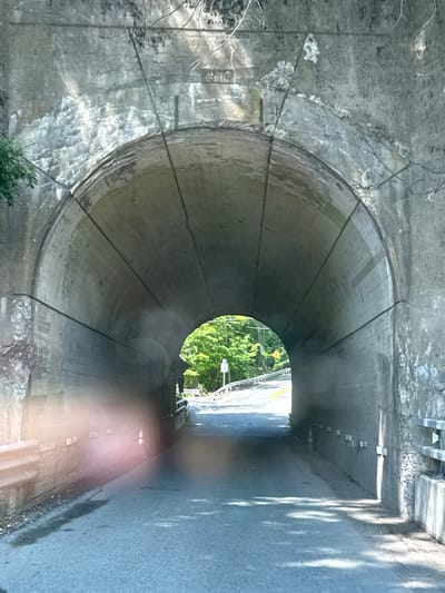 The image depicts the interior of a concrete tunnel, likely a road tunnel given the asphalt surface visible on the right. The tunnel has a circular or arched cross-section and appears to be constructed from segmented concrete panels. The walls are rough and weathered, with visible cracks and some white staining. Thin wires or cables run along the interior surface of the tunnel.
At the far end of the tunnel, a bright opening reveals a scene bathed in daylight. Sunlight filters through lush green trees, suggesting a time of day when the sun is high, likely late morning or afternoon. The weather appears to be clear and sunny, as indicated by the bright light and distinct shadows cast on the tunnel's exterior road surface.
The road surface within the tunnel is dark and appears wet in places, with some debris along the edges. Small, regularly spaced rectangular objects, possibly lights or markers, are embedded in the wall along the right side of the tunnel. There are no people visible in the image. The overall impression is one of a functional, somewhat aged piece of infrastructure set within a natural, green environment. The scene evokes a sense of transition between the enclosed darkness of the tunnel and the open, bright world outside. No visible text is present in the image.