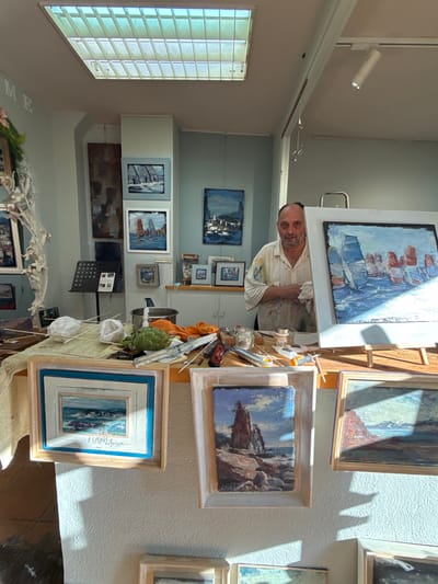 This media file captures an artist in their studio, surrounded by numerous paintings. The studio appears to be a bright and airy space, with sunlight streaming in from a window with blinds on the left side of the frame. The artist, a man with a beard and wearing a paint-splattered shirt, is actively engaged with a canvas on an easel, holding a rag in his hand.
The walls are adorned with a variety of framed artwork, primarily landscapes and seascapes. Several paintings depict boats in water, while others showcase rocky coastal scenes and abstract compositions. The overall impression is one of a working artist's studio, filled with creative output.
Notable details include the presence of art supplies scattered on a table next to the easel, such as brushes, jars, and tubes of paint. A metal stand with sheet music is also visible, suggesting the artist might also be a musician or that music plays a role in their creative process. A head of artichoke sits in a basket, adding a touch of still life to the scene.
The lighting suggests it is daytime, with strong shadows cast by the sunlight. The location context of Tossa De Mar, Spain, implies a potentially Mediterranean setting, which might be reflected in the coastal themes of the artwork. The artist's focused expression and the organized chaos of the studio convey a sense of dedication and passion for their craft. While no text is clearly legible on the artwork or within the scene, the overall atmosphere is one of artistic creation and inspiration.