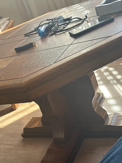This image shows a close-up view of a wooden table with a tiled surface. On the table, there are a pair of eyeglasses, a small blue flashlight, a black object with a gold tip (possibly a pen or a lighter), and a smartphone. The table is a light brown color with a textured surface, and it appears to be a dining or coffee table. Sunlight is streaming in from the side, casting shadows on the table and the floor, which is made of wood. The overall setting suggests an indoor environment. This image was taken in the city of Yeghvard, Armenia.