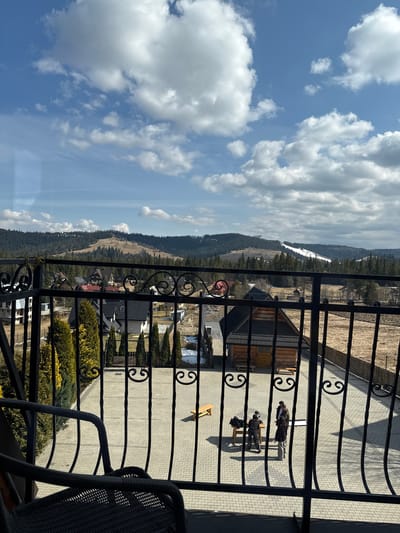 The image displays an elevated outdoor scene from a balcony overlooking a village and mountainous landscape in Murzasichle, Poland. In the foreground, a dark, ornate wrought-iron balcony railing with scrollwork is prominent, partially obscuring the view. The armrest and seat of a dark woven chair are visible in the lower left.
Below the balcony, a light-colored paved courtyard, constructed of interlocking rectangular pavers, extends into the midground. On this paved surface, three individuals are present. One person, dressed in dark clothing, kneels or bends over a light wooden table. Adjacent to this individual and the table, a camera on a tripod is set up, facing the table. A second person, wearing light pants and a dark jacket, stands facing the first individual and appears to hold a white rectangular panel, likely a reflector. A third person, clad in a long dark coat, stands slightly to the right, observing the activity. A second, simpler wooden bench is situated further back on the paved area to the left.
The courtyard is bordered on the left by a line of tall, slender evergreen trees. To the right, a rustic wooden building with a dark roof is visible, beyond which lies a fenced section of dry, fallow land with remnants of snow.
Further into the midground, the village of Murzasichle is visible, characterized by houses with dark roofs and light-colored or wooden facades, nestled among trees. Remnants of snow are scattered on the ground between properties.
The background consists of rolling hills and mountains covered in dense evergreen forests. A distinct white strip of snow, indicative of a ski slope, is visible on a distant hill to the right. The sky is bright blue with numerous scattered white cumulus clouds, and direct sunlight casts shadows on the paved courtyard.