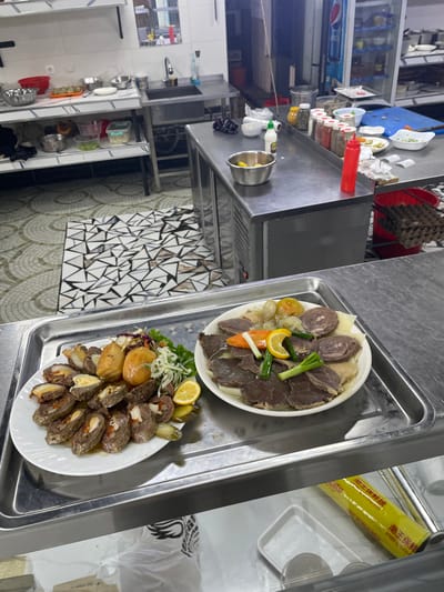 This image depicts a commercial kitchen scene in Chinabad, Uzbekistan. In the foreground, two large oval white platters of prepared food are presented on a rectangular silver serving tray, positioned on a lower-level stainless steel counter. The platter on the left contains numerous slices of a meat roll or sausage with a visible yellow interior and red elements, accompanied by small roasted potatoes, a fresh green and red cabbage salad, small pickled gherkins, and lemon wedges. The platter on the right displays slices of dark meat, consistent with horse meat (kazy/karta), arranged over boiled potatoes and onions, garnished with julienned carrots, sliced green onions, and lemon wedges.
Behind these dishes, a central stainless steel refrigerated counter is visible. On its surface, a silver bowl of lemon wedges, a white squeeze bottle, a row of approximately seven spice shakers, a red squeeze bottle, and several small white bowls containing garnishes or ingredients are present. Further to the right, a clear glass-front refrigerator with "PEPSI" branding is visible, along with a red plastic crate filled with brown egg cartons on the floor. To the left, a stainless steel counter features a sink with a faucet, above which are shelves holding various metal and plastic bowls. The floor in the foreground exhibits a distinctive geometric mosaic pattern of white, grey, and black triangles, transitioning to a lighter, circular tessellated pattern further into the background. No individuals are present in the scene. Below the main serving tray, a roll of yellow plastic wrap with Chinese characters is partially visible, alongside a white plastic bag and a white plastic tray.