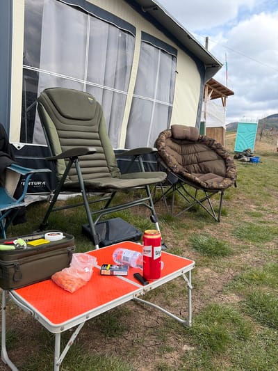 This image captures an outdoor scene within the specified city of Cherna Gora, Bulgaria, under an overcast sky. In the foreground, an unoccupied green, padded camping chair is positioned to the left of a small, orange-topped, silver-legged folding table. On the table, from left to right, are an open clear plastic bag containing multiple orange, spherical food items; a blue and white cigarette pack labeled "Marlboro Gold"; a black utility knife or lighter; a clear plastic water bottle lying on its side; and a red beverage can with white lettering, consistent with a beer brand. An olive green soft-sided utility bag, containing a white spool and other small items, rests on the ground to the left of the table. To the right of the green chair, an unoccupied brown, camouflaged, padded, rounded camping chair is visible. Behind these items, a large, light beige canvas tent or caravan awning with clear plastic windows forms the backdrop. In the mid-ground, to the right, a partially covered wooden structure and a light blue portable toilet are present. Distant rolling hills are visible under the cloudy sky, and a Bulgarian flag is faintly discernible on a pole behind the wooden structure. No individuals are visible in the scene.