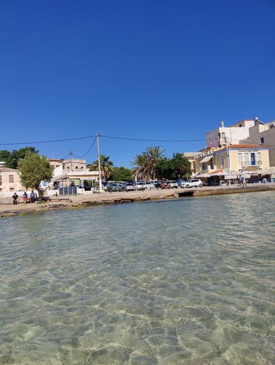 The image shows a clear, sunny day with a bright blue sky over a waterfront scene in Aegina, Greece. The foreground is dominated by calm, transparent turquoise water, revealing the sandy bottom beneath. Along the shore, a paved walkway runs parallel to a row of buildings.

On the left side of the walkway, three men are seated on a low stone wall, facing the water. Further along, a small green tree provides shade. Adjacent to the tree, a building with light-colored walls and brown shutters is visible.

A white awning stretches across the front of a building that appears to be a restaurant or cafe. Underneath the awning, tables and chairs are set up. The text "ΚΑΗΛΥΡΓΙΝΑ" is visible on a sign above the awning.

To the right of this establishment, more buildings line the waterfront. One prominent yellow building has a blue balcony door and white window frames. Several cars are parked along the street behind the buildings, indicating it is likely a public area or a parking lot. Palm trees are scattered throughout the scene, adding to the Mediterranean atmosphere.

The overall impression is a tranquil daytime scene at a seaside town, likely during warm weather, given the clear sky and the presence of people enjoying the outdoors by the water. The sunlight casts bright reflections on the water and illuminates the colorful buildings.