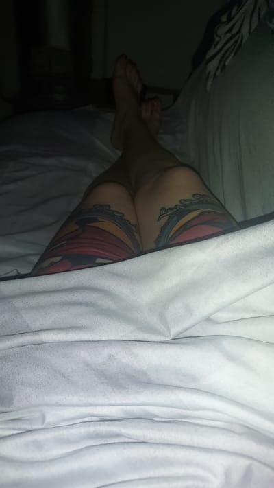 The image shows a person's legs with tattoos on them, lying on a white bed. The lighting is dim, and the background is out of focus, showing a dark room. The person's feet are visible, with painted toenails. The tattoos appear to be colorful and detailed, with designs resembling mouths and flames. The scene is intimate and personal, suggesting a private setting. There is no visible indication of the location being Kogon City, Uzbekistan.