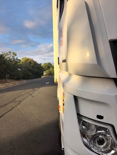 A white truck is parked on the side of a road, with its front right visible. The truck has a logo on its side and the number "B45" on the hood. The road is asphalt and has a white line marking. There are trees and bushes on the left side of the road. The sky is blue with some clouds. The scene is likely in Saint-Augustin-des-Bois, France, judging by the presence of a yellow and black sign on the truck, which indicates it is an overloaded vehicle, a common sight in European countries.