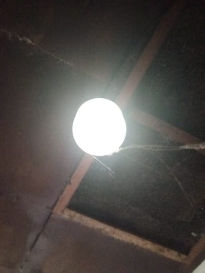 The image depicts an upward-angled view of an interior ceiling located in Keo Kouang, Laos. A single, intensely bright, spherical white light source, presumed to be an incandescent or LED bulb, occupies the upper-center of the frame, emitting significant glare. This light illuminates a portion of the dark brown ceiling structure, which features exposed wooden beams forming an angular, possibly structural, framework. The areas between the beams are covered with dark, textured material, potentially panels or netting, showing signs of dust and debris accumulation. Multiple strands of light-colored cobwebs are visible, extending from the vicinity of the light source towards the upper-right and lower-right sections of the ceiling, adhering to both the light fixture and the ceiling material. A dark electrical cable is visible running horizontally along a beam in the lower-left quadrant of the image. No people, subjects, or actions are present.