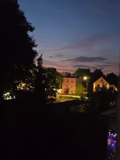A low-angle, long-exposure photograph captures a twilight scene in Offenbach am Main, Germany, with the sky transitioning from a deep blue to hues of pink and purple. Silhouetted trees dominate the foreground, their dark foliage obscuring much of the lower portion of the frame. Behind the trees, a cluster of buildings is visible.

To the left, a multi-story building with illuminated windows suggests residential or office spaces. In the center, a light pink building with square windows is illuminated, with a street lamp casting a bright glow on the right side of it. To the right of the pink building, a yellow house is also lit, with its windows and the facade warmly illuminated. Trees and foliage surround these structures, some of which are illuminated by the building lights.

In the background, a construction crane is visible against the dusky sky. The overall impression is that of a quiet residential area at dusk, with the artificial lights of the buildings and street lamps beginning to punctuate the fading natural light. The scene is calm and still, with no human figures or discernible activity visible.