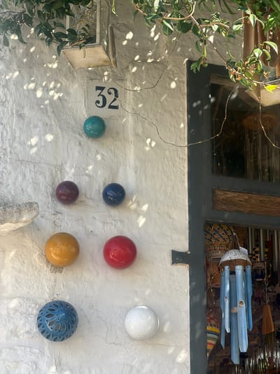 An outdoor medium shot captures a textured white wall with various decorative spherical objects and a house number. The house number "32" is prominently displayed in black on a white square background. Below the number, a teal sphere is mounted on the wall. Further down, in a diagonal line, are a maroon sphere, a navy blue sphere, a yellow sphere, and a red sphere. At the bottom left, a blue patterned spherical object is visible, and to its right, a white spherical object. The wall is illuminated by dappled sunlight, casting shadows from overhead foliage and a string of small lights. To the right of the wall, a dark-colored doorway or window frame is partially visible, with a wooden beam across it and a glimpse of wind chimes and other decorative items hanging behind. The scene appears to be an exterior wall of a building in Alberobello, Italy, possibly a home or shop, characterized by its traditional whitewashed architecture and decorative elements.