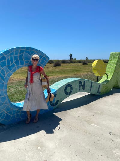 A blonde woman stands in front of a large, brightly colored mosaic sculpture in Conil de la Frontera, Spain. The sculpture appears to be letters spelling out "CONIL" with a circular yellow element on top. The woman is wearing sunglasses, a light-colored tiered dress, a red scarf around her neck, and sandals. She is holding a straw bag over her shoulder and a straw hat in her right hand. The background shows a clear blue sky, green grass, and some sparse vegetation and palm trees. The sculpture is made of blue, green, and yellow tiles. The ground is paved with concrete. The scene is well-lit by sunlight, casting a shadow from the woman.