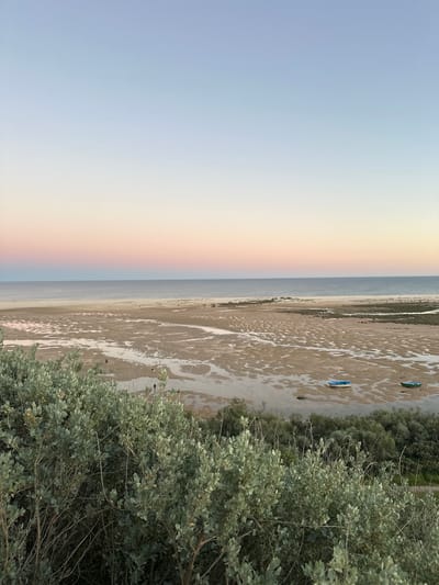 The image shows a beach landscape, possibly in Vila Real de Santo António, Portugal. The view is from an elevated position, partially obscured by a bush with gray-green leaves. To the left is the sky, transitioning from light blue to a soft orange-yellow as it nears the horizon. A pale rainbow is visible near the center. The sea meets a sandy beach. A large mudflat is prominent, interspersed with shallow pools of water. Two small, blue boats are visible on the mudflat. A strip of vegetation borders the mudflat.