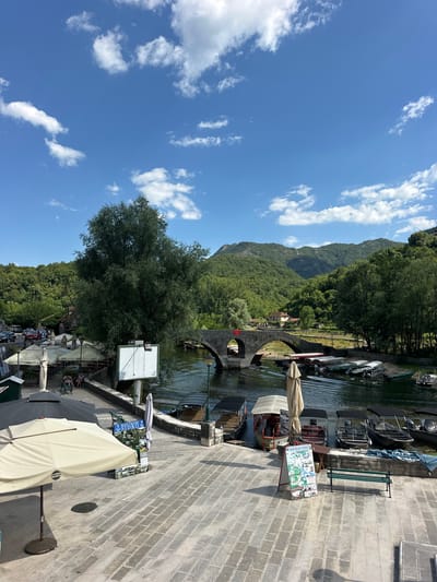This is a media file depicting a scenic riverfront view in Rijeka Crnojevića, Montenegro, on a bright, sunny day with a clear blue sky and scattered white clouds. The scene is characterized by a tranquil river, lined with boats docked along its banks. Several boats, varying in size and design, are visible, some appearing to be fishing boats or small passenger vessels.
In the foreground, a paved walkway runs along the river, adorned with outdoor seating areas under numerous umbrellas, suggesting a lively restaurant or cafe setting. Some of these umbrellas are tan, while others are dark grey. One tan umbrella prominently displays a "PEPSI" logo. There are a few people seated at tables and on benches, enjoying the day. A sign with multiple panels, displaying images and text, stands near the walkway, possibly advertising local attractions or services.
Further into the scene, an old stone bridge with multiple arches spans the river, adding a historic element to the landscape. Beyond the bridge, lush green hills and mountains covered in dense forest rise majestically, creating a picturesque backdrop. A few buildings are visible nestled amongst the trees on the hillside. The water of the river reflects the blue sky and surrounding greenery, contributing to the serene atmosphere. The time of day appears to be midday, judging by the strong sunlight and the shadows cast. The overall mood is one of peaceful relaxation and natural beauty.