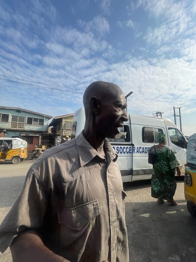 The image captures a street scene in Akowonjo, Nigeria, under a partly cloudy sky. In the foreground, a bald Black man is shown from the chest up, wearing a light brown button-down shirt with two chest pockets. He is looking to his right with his mouth open as if speaking. Behind him, a white van with "SOCCER ACADEMY" written in blue letters on its side is parked. To the left of the van, a yellow auto-rickshaw is visible. Further in the background, several multi-story buildings with windows and balconies line the street. A woman in a green and black patterned dress and headscarf walks away from the van towards the right side of the frame. The ground is paved with asphalt, and power lines are visible crisscrossing the sky.