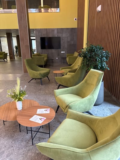 The image depicts an indoor waiting area, likely in a commercial or office building. The space features several modern, olive-green upholstered chairs with black metal legs. Two small, oval-shaped wooden coffee tables are positioned in the foreground. One table holds a white vase with a dried floral arrangement, and the other has two printed flyers on it. In the background, more green chairs are visible, along with a television mounted on a dark grey tiled wall. The room is decorated with a bright yellow wall and a wooden slatted wall panel. The flooring is a speckled grey tile. There are no people visible in the image. The location is Kholmy, Russia.