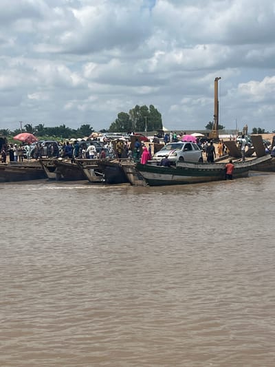 The image depicts a ferry crossing point in Nemnei, Nigeria, with multiple wooden boats laden with people and vehicles on a muddy river. The sky is overcast with dramatic clouds, suggesting a humid, tropical climate. On the shore, a construction crane is visible in the background, hinting at ongoing development or infrastructure projects. People are seen disembarking from the boats and moving towards the shore, while others wait to board. A silver car is partially loaded onto one of the boats, indicating the transport of vehicles across the river. The water is a murky brown, characteristic of a river with a high sediment load. The scene conveys a sense of busyness and activity at a vital transportation hub within the community.
